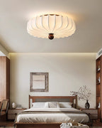 A Clérachic ceiling light lit above a calm bedroom, with a framed print, a wooden bed and shelves in the background.