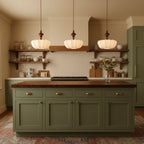 Three Clérachic pendants in a row above a green kitchen island, each shade glowing softly beside open shelving.
