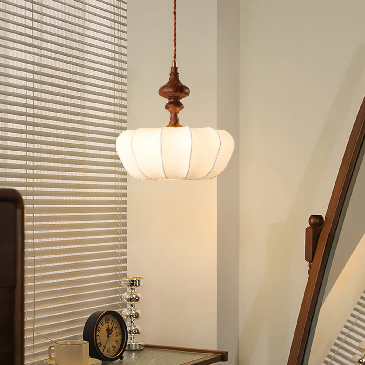 Ribbed white silk shade of a Clérachic pendant glowing in a corner beside slatted blinds, with a small clock below.