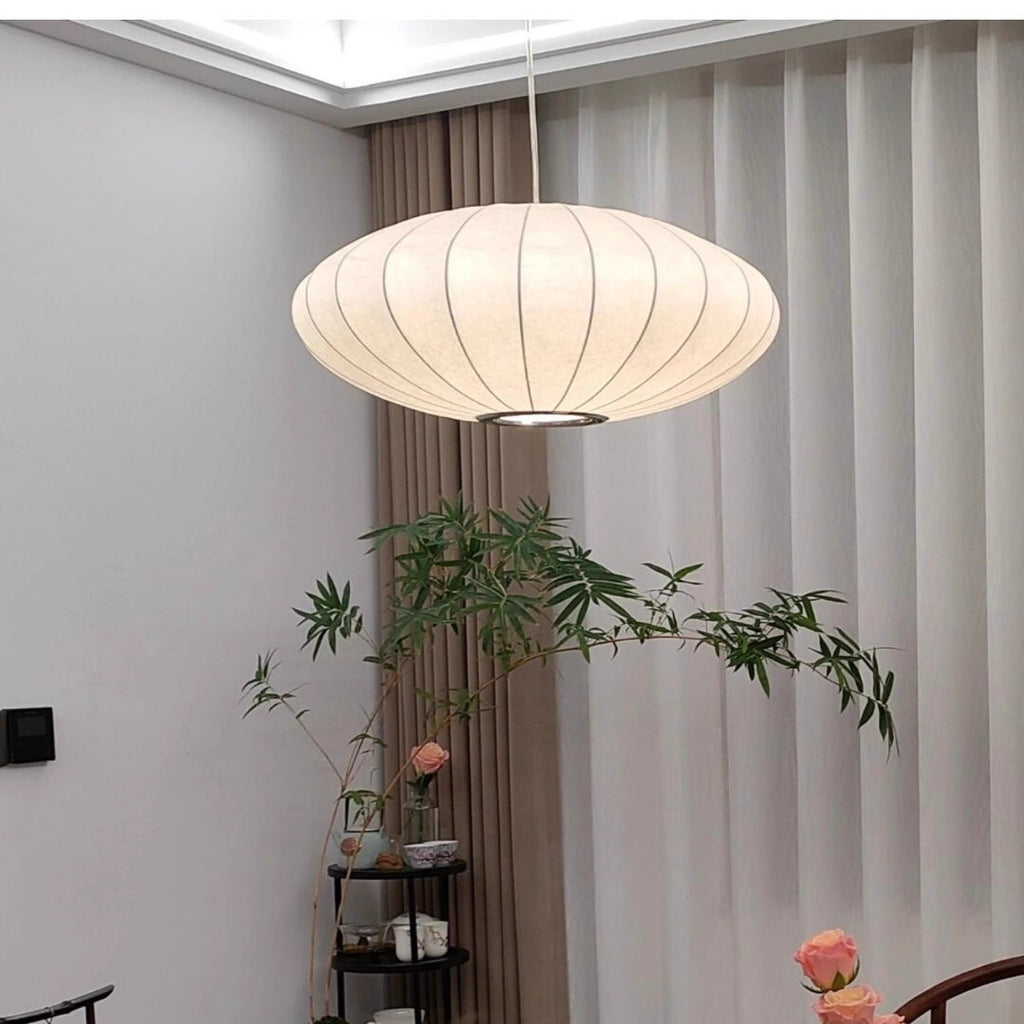 Clérachic saucer pendant lamp hanging unlit in a daylit room beside sheer curtains, the metal ring around its base opening catching the light above a bamboo plant.