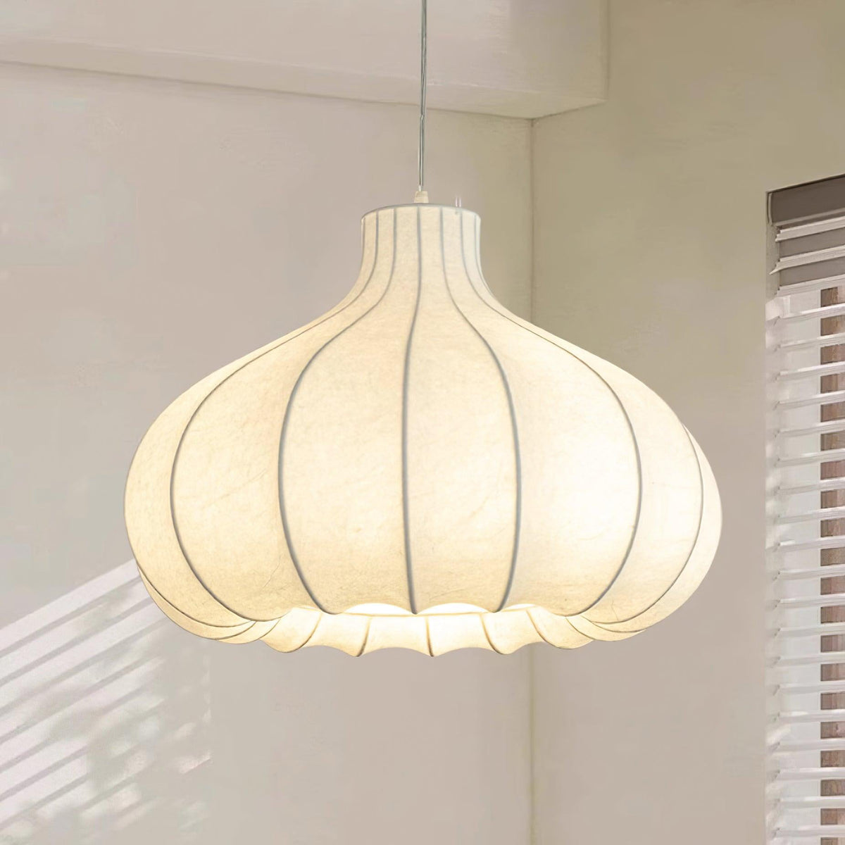 Clérachic garlic-bulb silk pendant light beside a window with white slatted blinds, daylight striping the wall behind the ribbed shade.