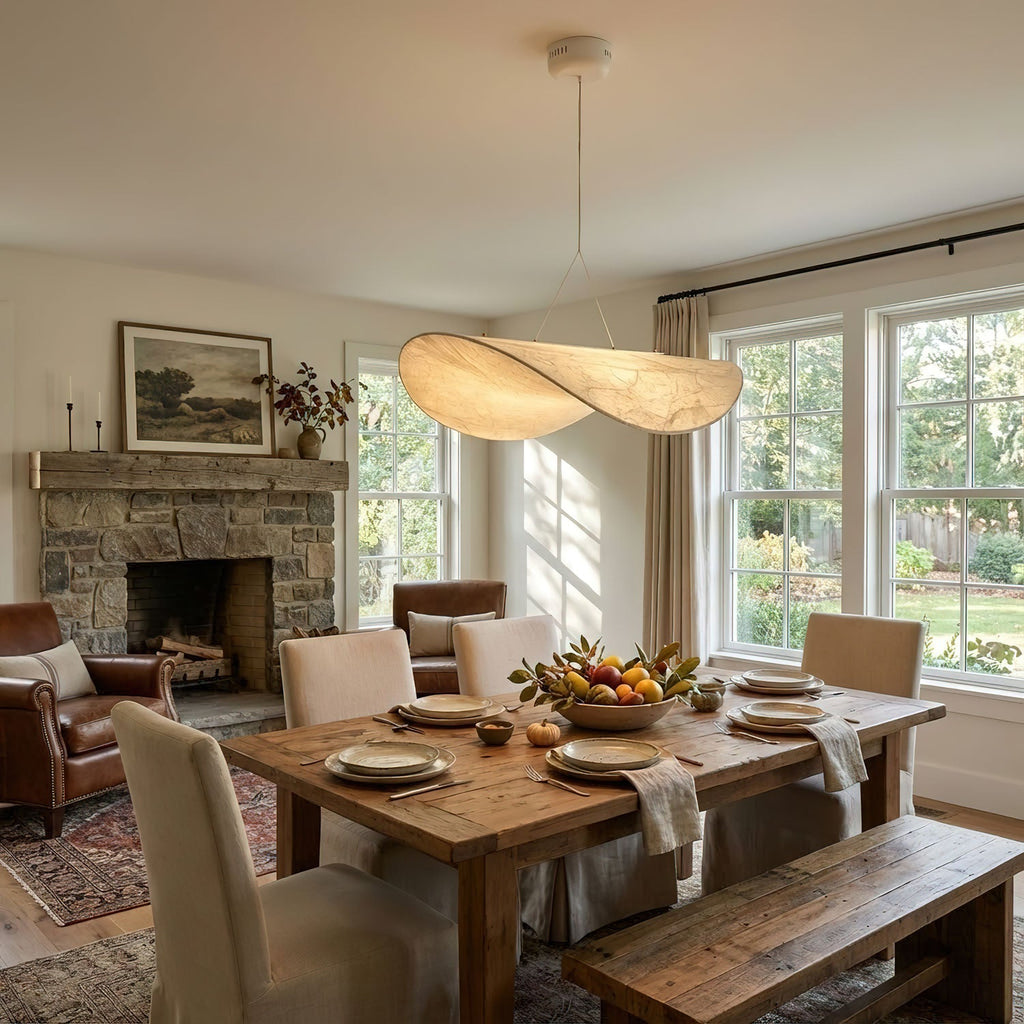 A twisted white pendant hangs over a rustic dining table and benches, with a stone fireplace and leather armchair beyond, from Clérachic.