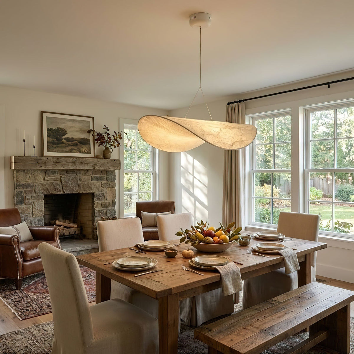 A twisted white pendant hangs over a rustic dining table and benches, with a stone fireplace and leather armchair beyond, from Clérachic.