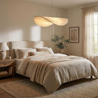 A twisted white pendant glows above a bed with oatmeal linen and a woven rug, beside a tall window with sheer curtains, from Clérachic.