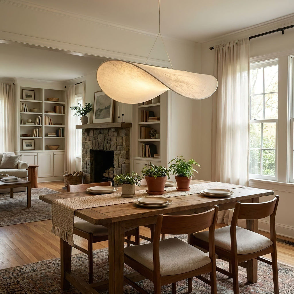 A white pendant with a twisted edge hangs above a wooden dining table with cushioned chairs, a stone fireplace and bookcases behind, from Clérachic.