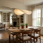 A white pendant with a twisted edge hangs above a wooden dining table with cushioned chairs, a stone fireplace and bookcases behind, from Clérachic.