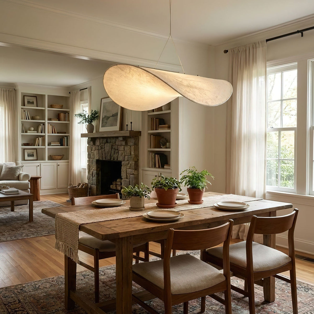 A white pendant with a twisted edge hangs above a wooden dining table with cushioned chairs, a stone fireplace and bookcases behind, from Clérachic.