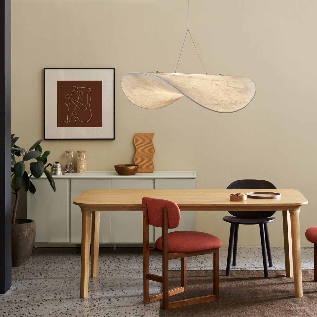 A twisted white pendant hangs over a pale oak dining table with a red-cushioned chair, beside a framed print and a low sideboard, from Clérachic.