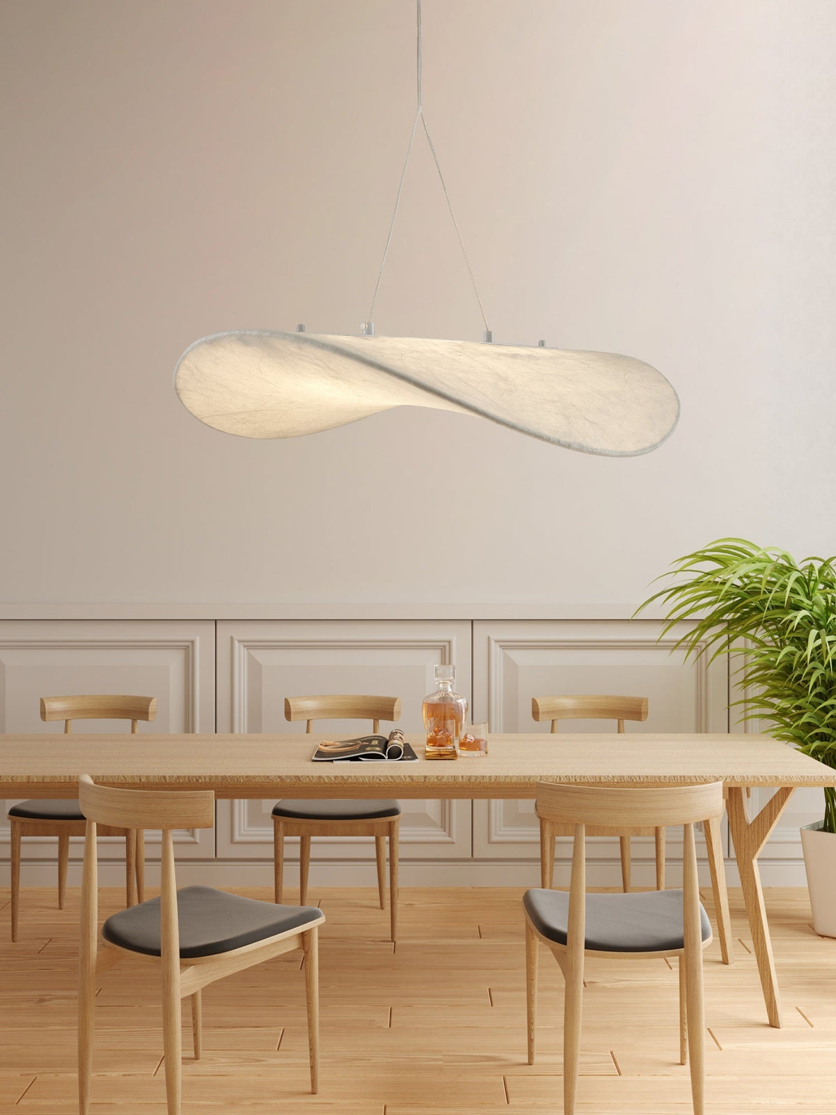 A twisted white pendant hangs above a long pale oak table with light chairs, in front of a panelled wall and a green plant, from Clérachic.