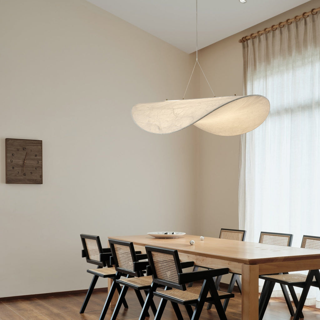 A twisted white pendant hangs in a tall, bright dining room with sheer curtains, a pale oak table and black cane-back chairs, from Clérachic.