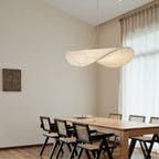 A twisted white pendant hangs in a tall, bright dining room with sheer curtains, a pale oak table and black cane-back chairs, from Clérachic.