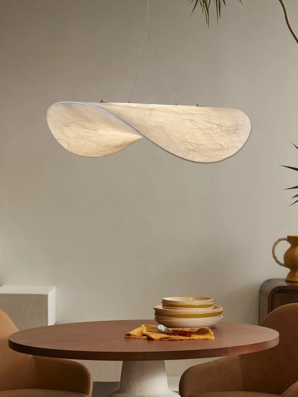A twisted white pendant hangs against a beige wall above a round walnut table set with stacked bowls, from Clérachic.