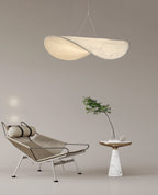 A twisted white pendant hangs in a taupe room above a wire lounge chair and a small marble side table with a green branch, from Clérachic.