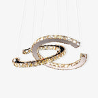 Two interlocking crescent rings of cognac-toned crystal on polished steel, a Clérachic pendant hung from fine cables on a white backdrop.