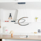 Distant view of the Clérachic crescent pendant, unlit, in a white room with shelves, a table lamp and a wooden counter.