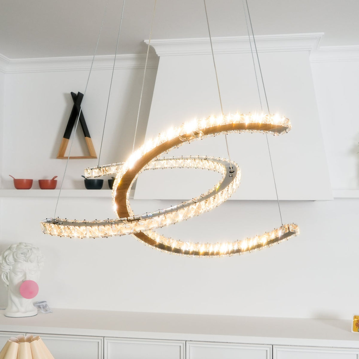 Clérachic pendant lit warm, its crystals glittering along both crescents, in front of a white shelving wall.