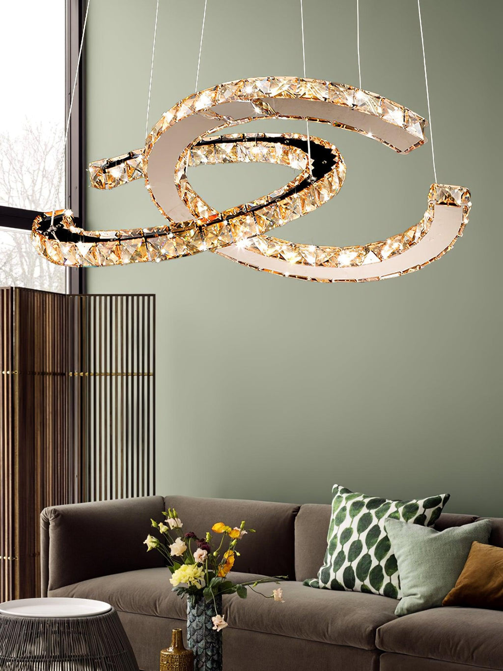 Cognac-toned Clérachic pendant lit beside a sage-green wall, with a brown sofa and a bunch of yellow flowers below.