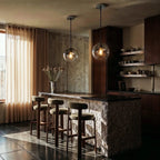 Clérachic glass sphere pendants light a stone kitchen island, paired above dark timber cabinetry and bar stools.