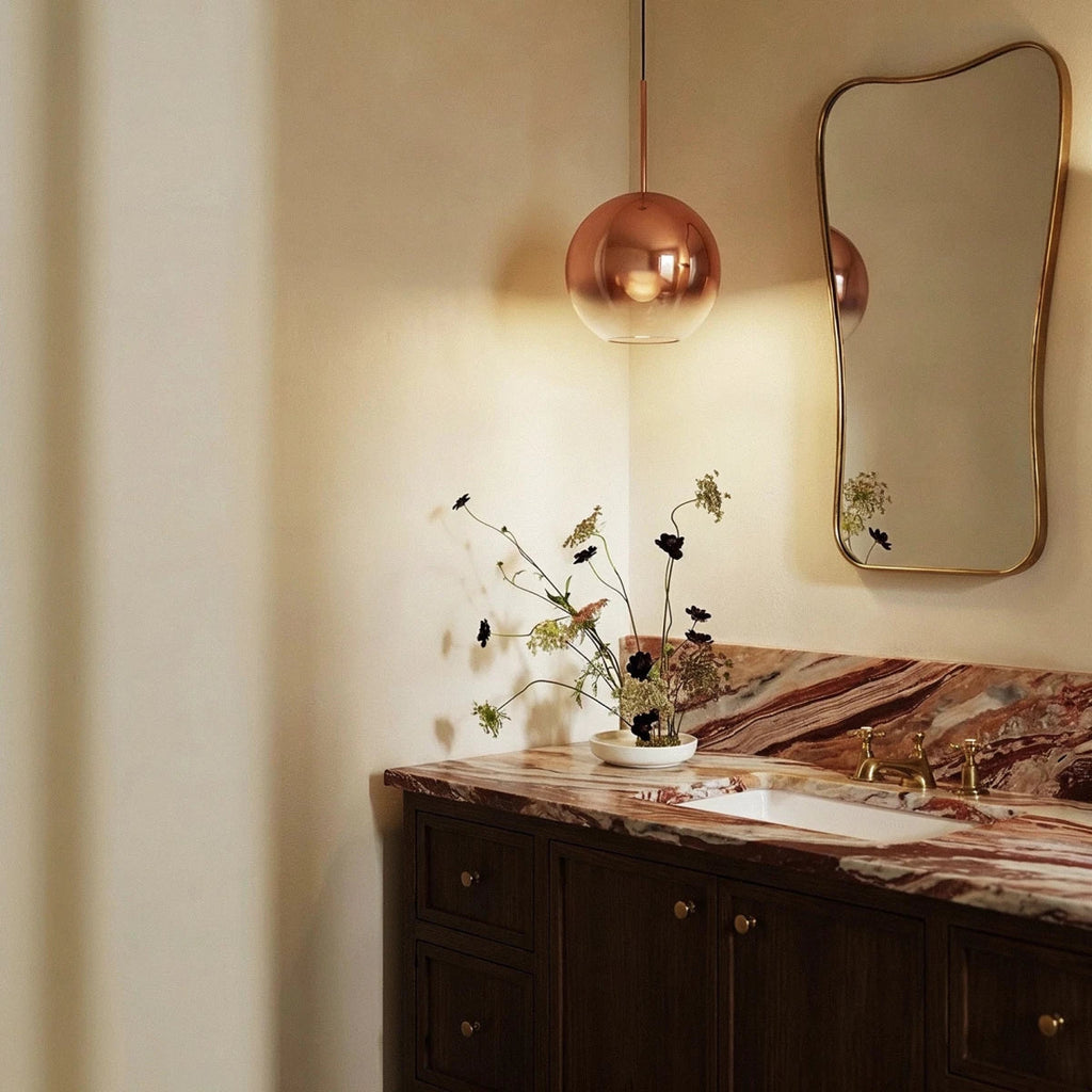 Clérachic rose gold glass sphere pendant hangs beside a bathroom vanity mirror above a veined stone counter.