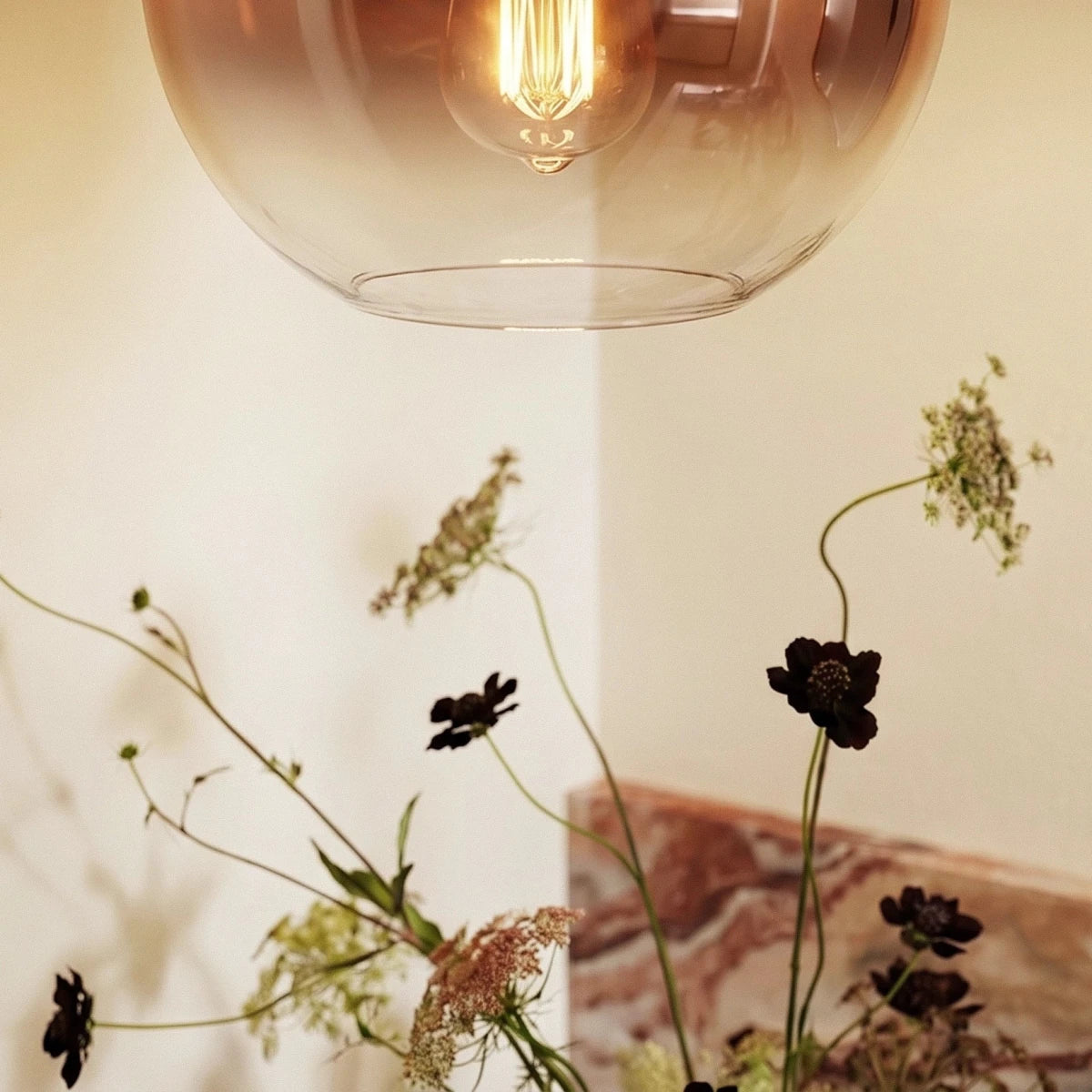 Clérachic close-up of the rose gold gradient glass sphere and glowing bulb, framed by dried flowers below.
