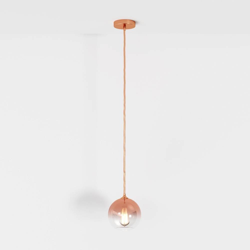 Clérachic rose gold pendant light hangs on a matching cord, glass sphere shade lit against a white backdrop.
