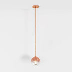 Clérachic rose gold pendant light hangs on a matching cord, glass sphere shade lit against a white backdrop.