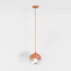 Clérachic rose gold pendant light drops from a matching ceiling canopy, glass sphere glowing softly on white.