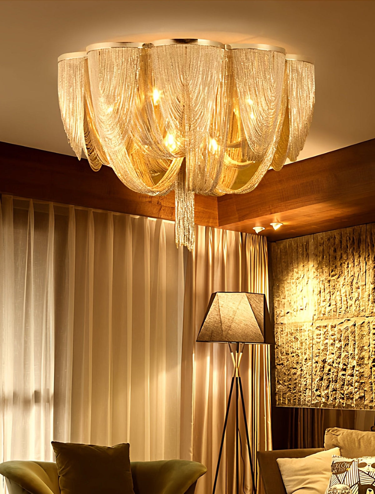 Gold-toned Clérachic chain ceiling light glowing warm in a room with honey-coloured curtains, a tripod lamp and a stone wall.
