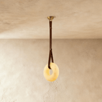 Clérachic alabaster ring pendant light glowing warm, hung from a brown leather strap and gold canopy on a limewash wall.