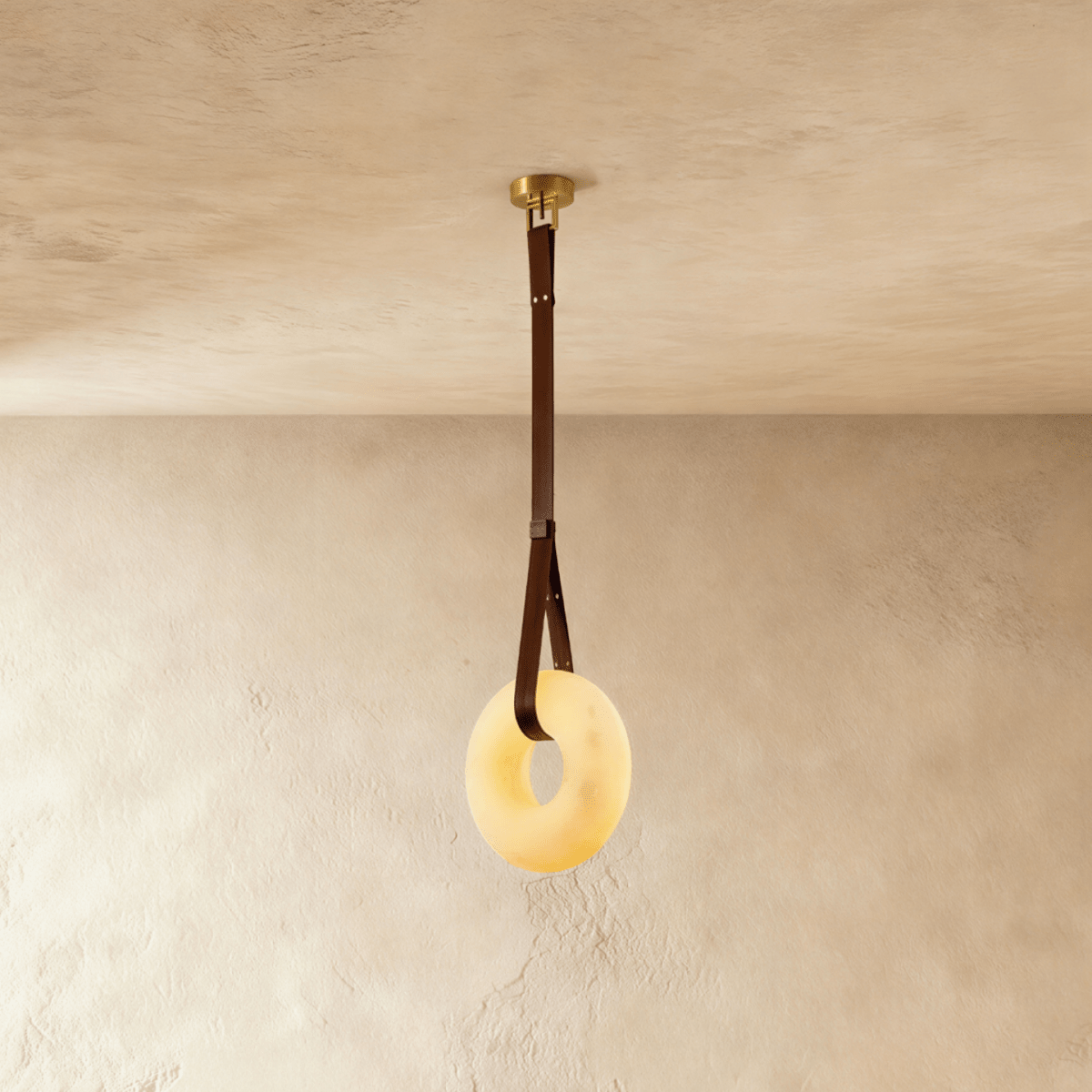 Clérachic alabaster ring pendant light glowing warm, hung from a brown leather strap and gold canopy on a limewash wall.