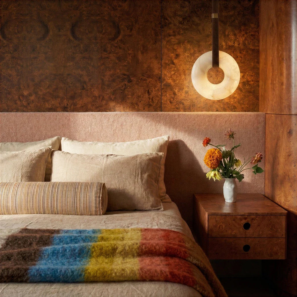 Clérachic alabaster ring pendant light glowing beside a bed, against dark burr wood panelling and a blush headboard.