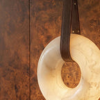 Detail of the Clérachic alabaster ring pendant light with stitched brown leather strap against warm burr wood panelling.