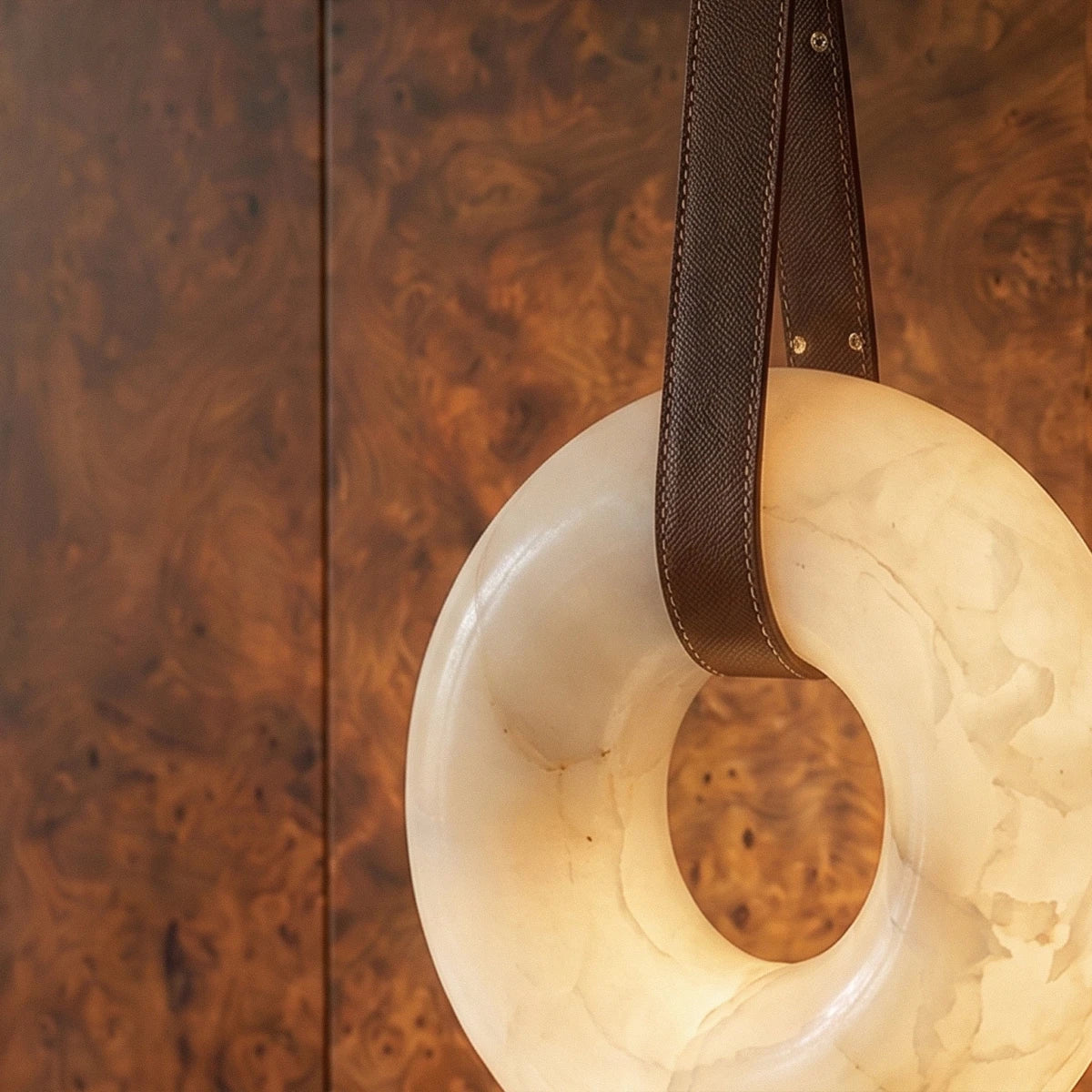 Detail of the Clérachic alabaster ring pendant light with stitched brown leather strap against warm burr wood panelling.