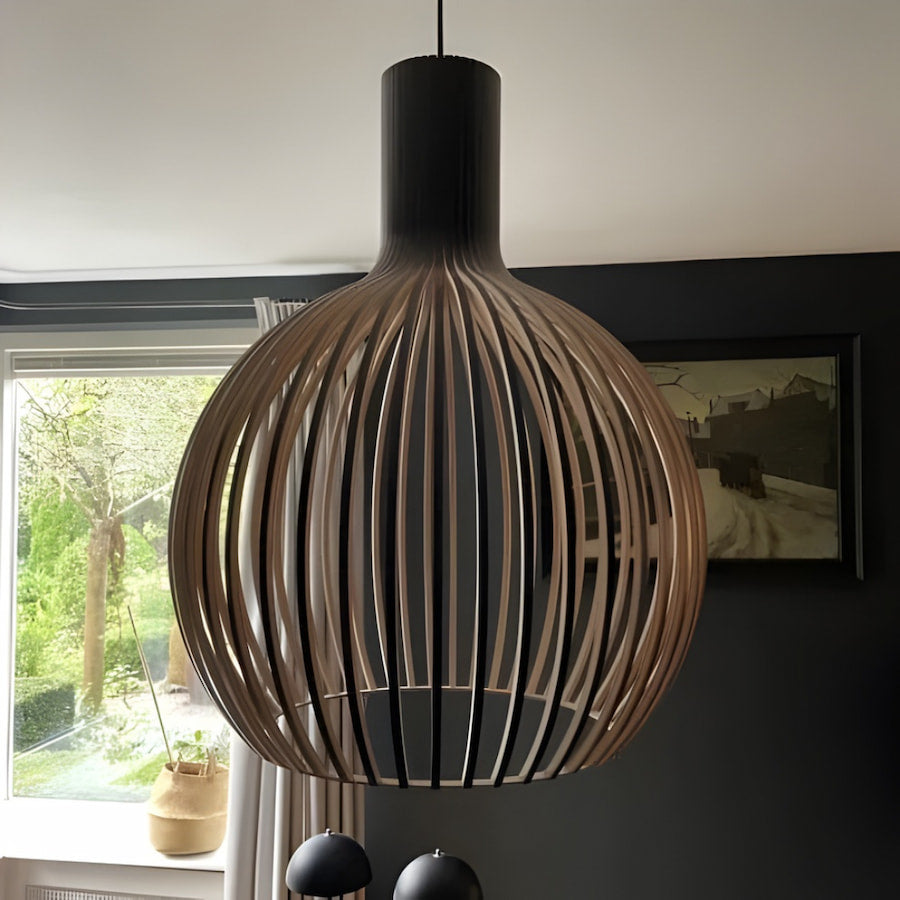 Clérachic wood-slat pendant light with a black collar and pale wood ribs, hanging unlit against a dark wall beside a window looking onto a garden.
