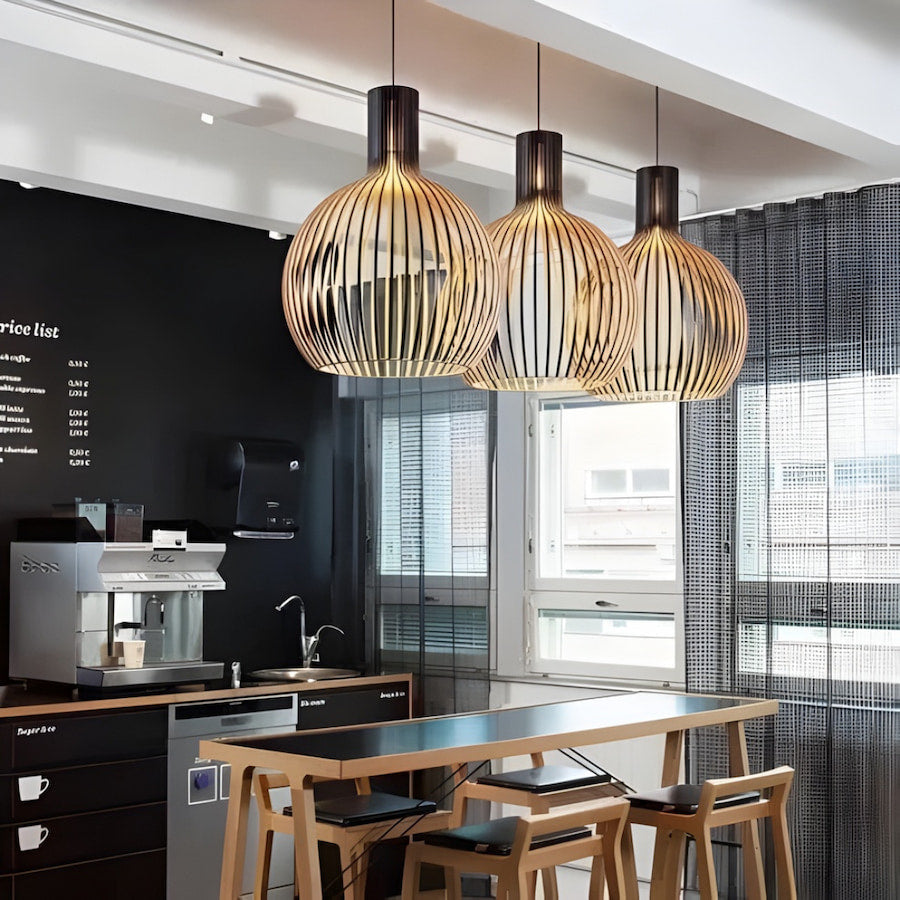 Clérachic wood-slat pendant lights with dark collars, three hung in a row above a bar-height table in a café with an espresso machine and a chalkboard price list.