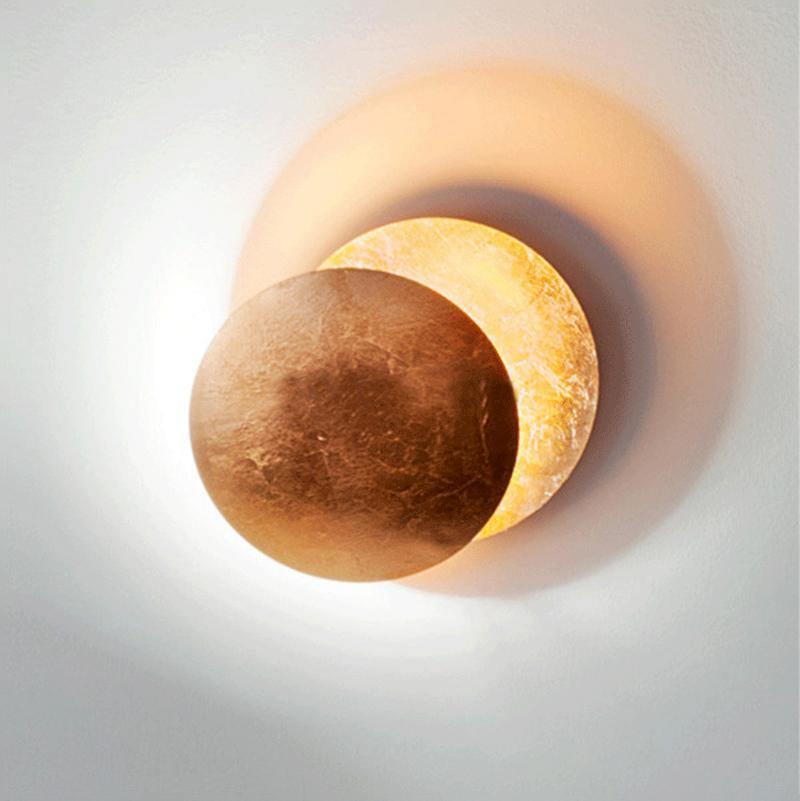 Lit Clérachic wall light on a pale wall, a bronze-brown front disc half covering a gold-leaf disc with a bright glow spilling around both.