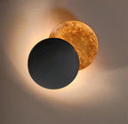 Clérachic wall light with a matt black front disc over a gold-leaf disc, glowing warmly against a soft taupe wall.