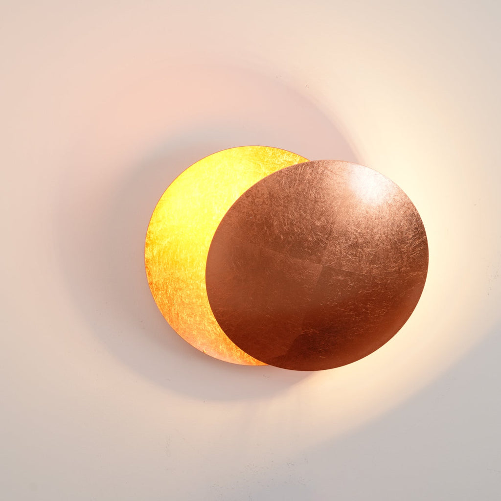 This Clérachic wall light lit at dusk, a copper-brown disc in front and a bright gold disc behind it, with a peach glow on the wall.