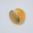 Unlit Clérachic wall light in gold, its front disc angled forward to reveal a deeper amber disc behind, on a pale grey wall.