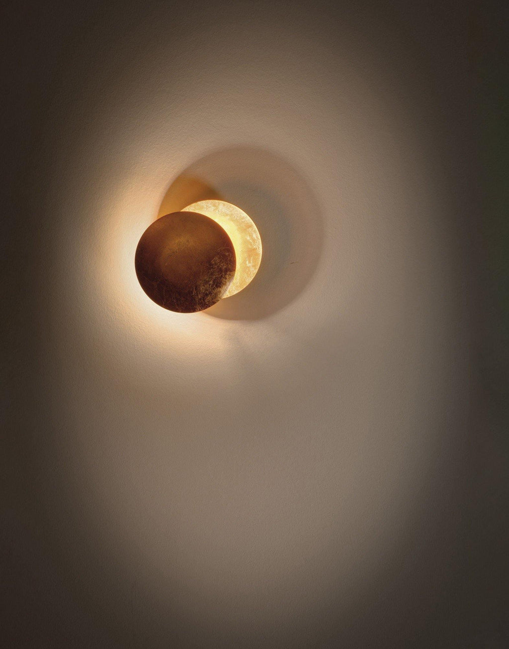 Narrow view of a lit Clérachic wall light on a dark taupe wall, a brown disc overlapping a gold one inside a wide soft glow.