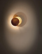 Narrow view of a lit Clérachic wall light on a dark taupe wall, a brown disc overlapping a gold one inside a wide soft glow.