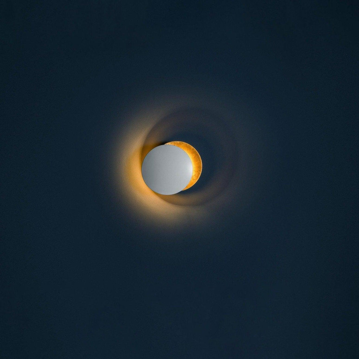 Small Clérachic wall light on a dark navy wall, a pale disc beside a gold one, with a faint amber light along the rim.