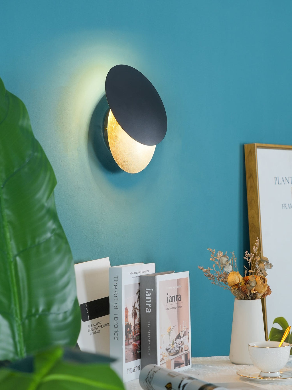 Clérachic wall light on a teal wall above books and a vase of dried flowers, its black disc tilted to show a lit gold disc behind.