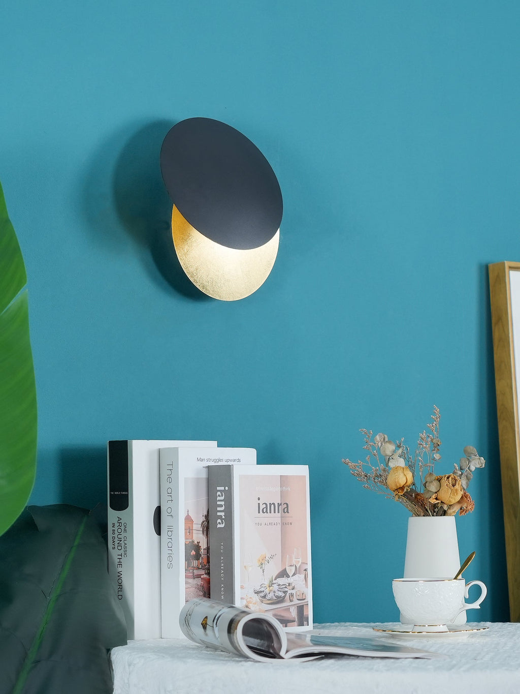 Black and gold Clérachic wall light on a teal wall, set above a stack of books, a white vase of dried flowers and a coffee cup.
