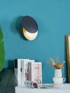 Black and gold Clérachic wall light on a teal wall, set above a stack of books, a white vase of dried flowers and a coffee cup.