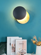 Closer view of this Clérachic wall light on a teal wall, a black disc overlapping a glowing gold disc above art books and dried flowers.