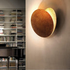 Clérachic wall light on a cream wall beside a full bookcase, a bronze front disc swung forward from a gleaming gold disc.