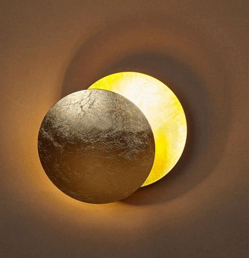 Lit Clérachic wall light on a warm brown wall, an antique-gold front disc partly covering a brighter gold disc that lights the wall.