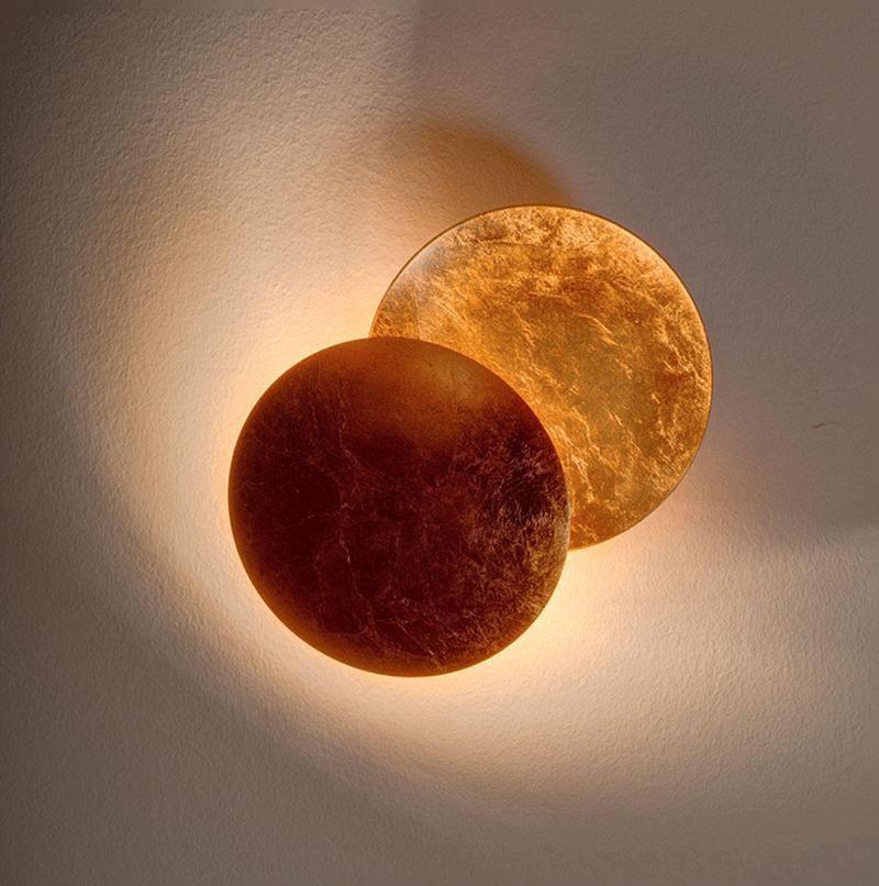 Large Clérachic wall light with a deep bronze front disc and a gold-leaf disc behind, lit softly against a grey-beige wall.