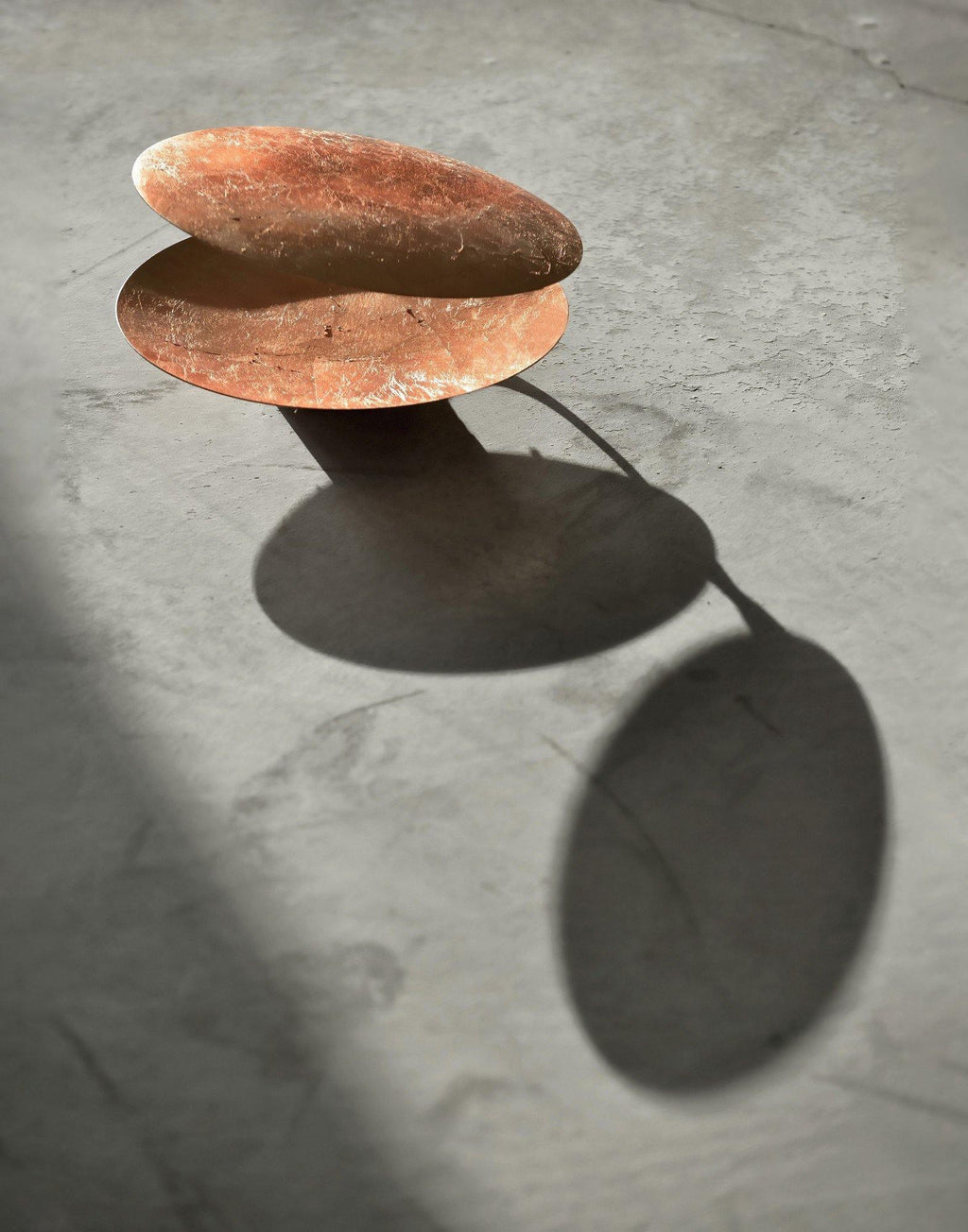 Copper-leaf discs from this Clérachic wall light standing on a concrete floor, their shadows stretching away in side light.